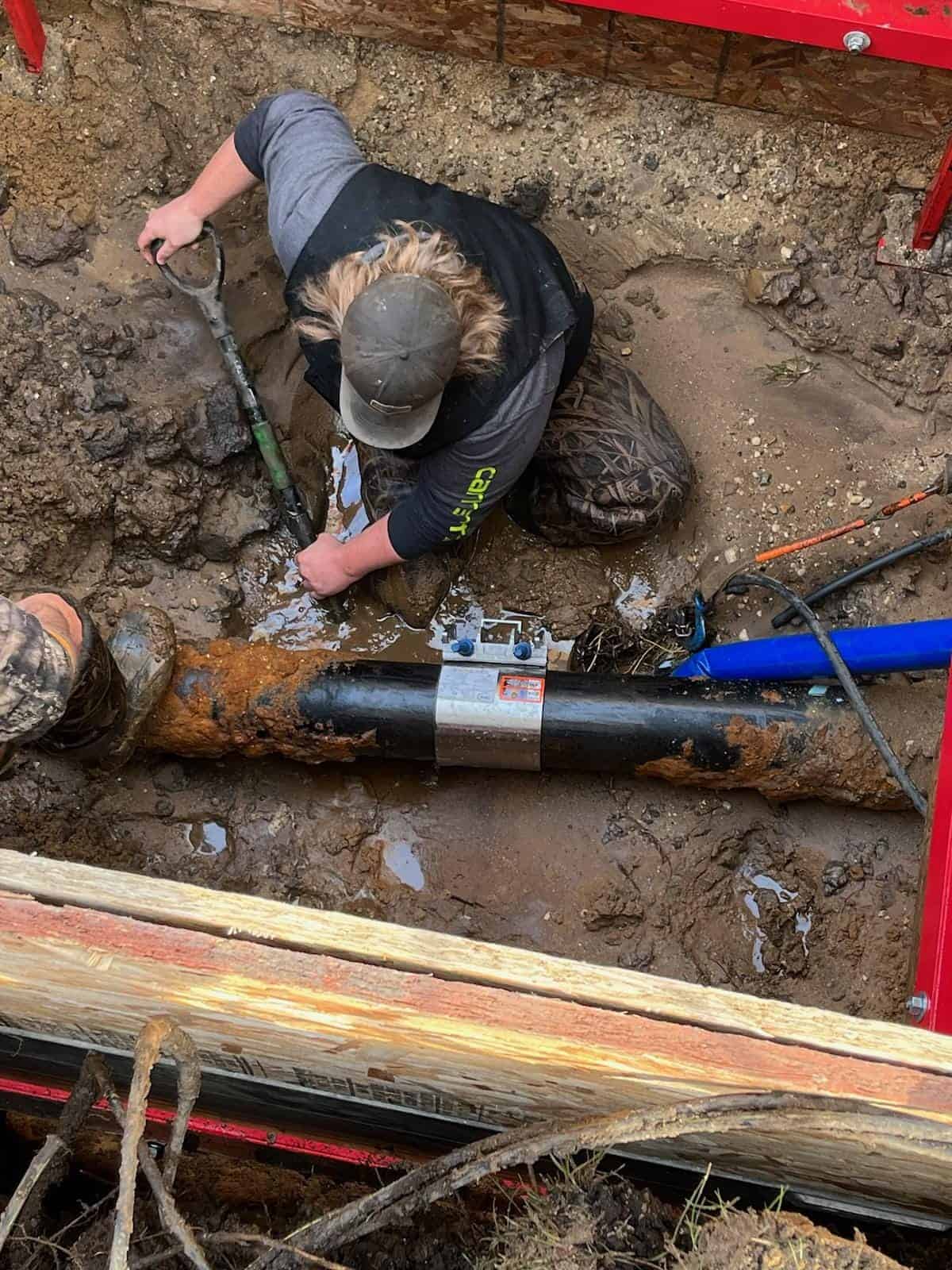 Worker repairing a pipe in a muddy trench.